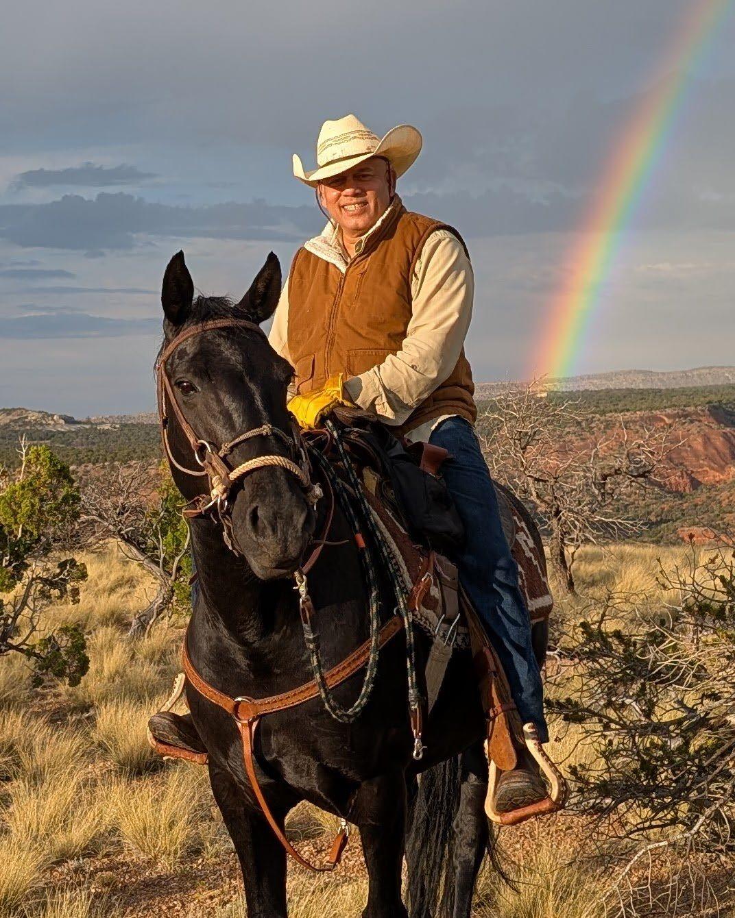 Paul D'Souza in a straw cowboy hat and tan vest sitting Rosa, a black mare in a braided bridle, on a juniper ridge in low evening sun with a rainbow falling behind him