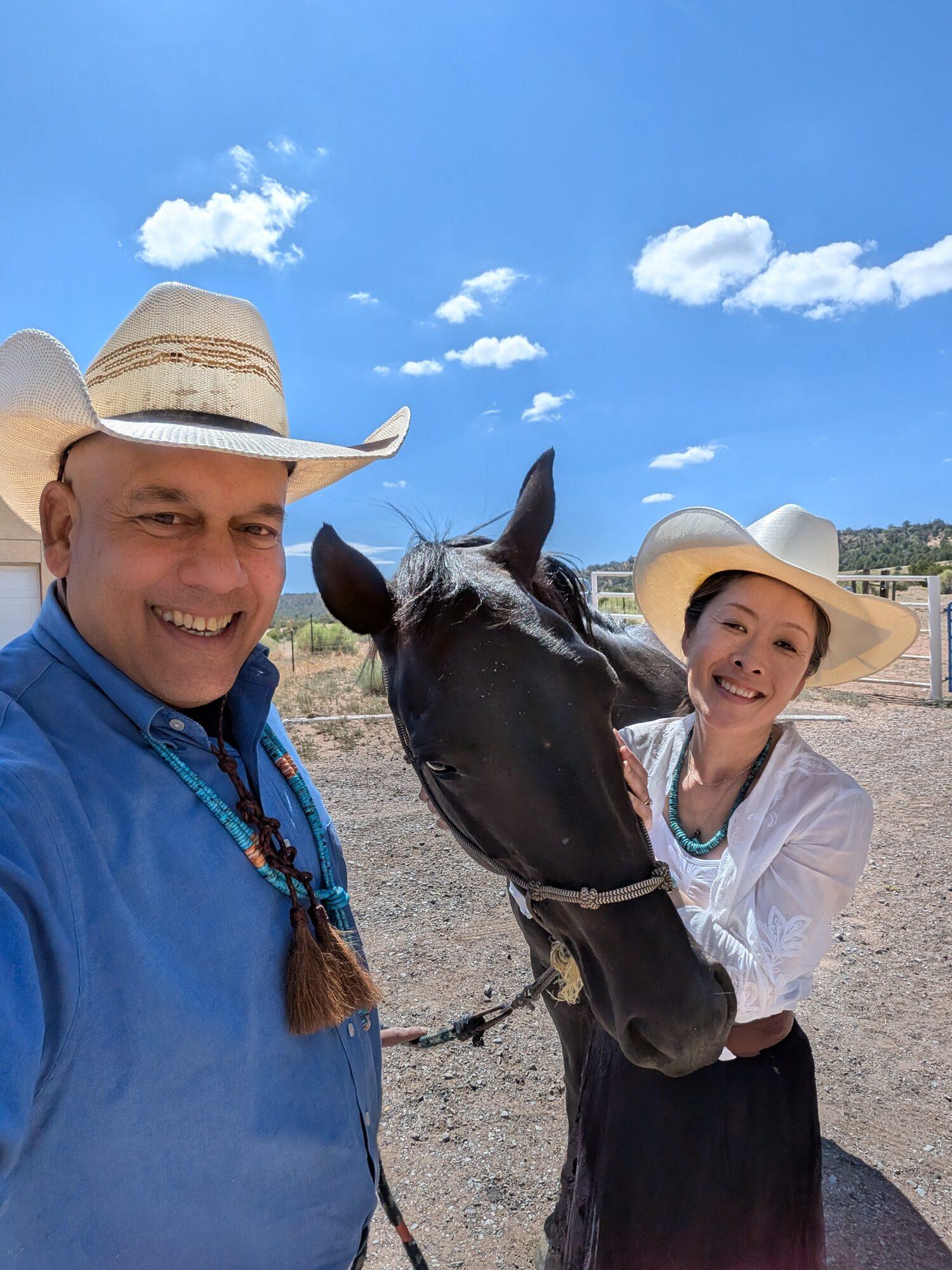Paul D'Souza and Shino in straw cowboy hats standing either side of Rosa, a black mare in a rope halter, at the ranch corral under a clear blue sky