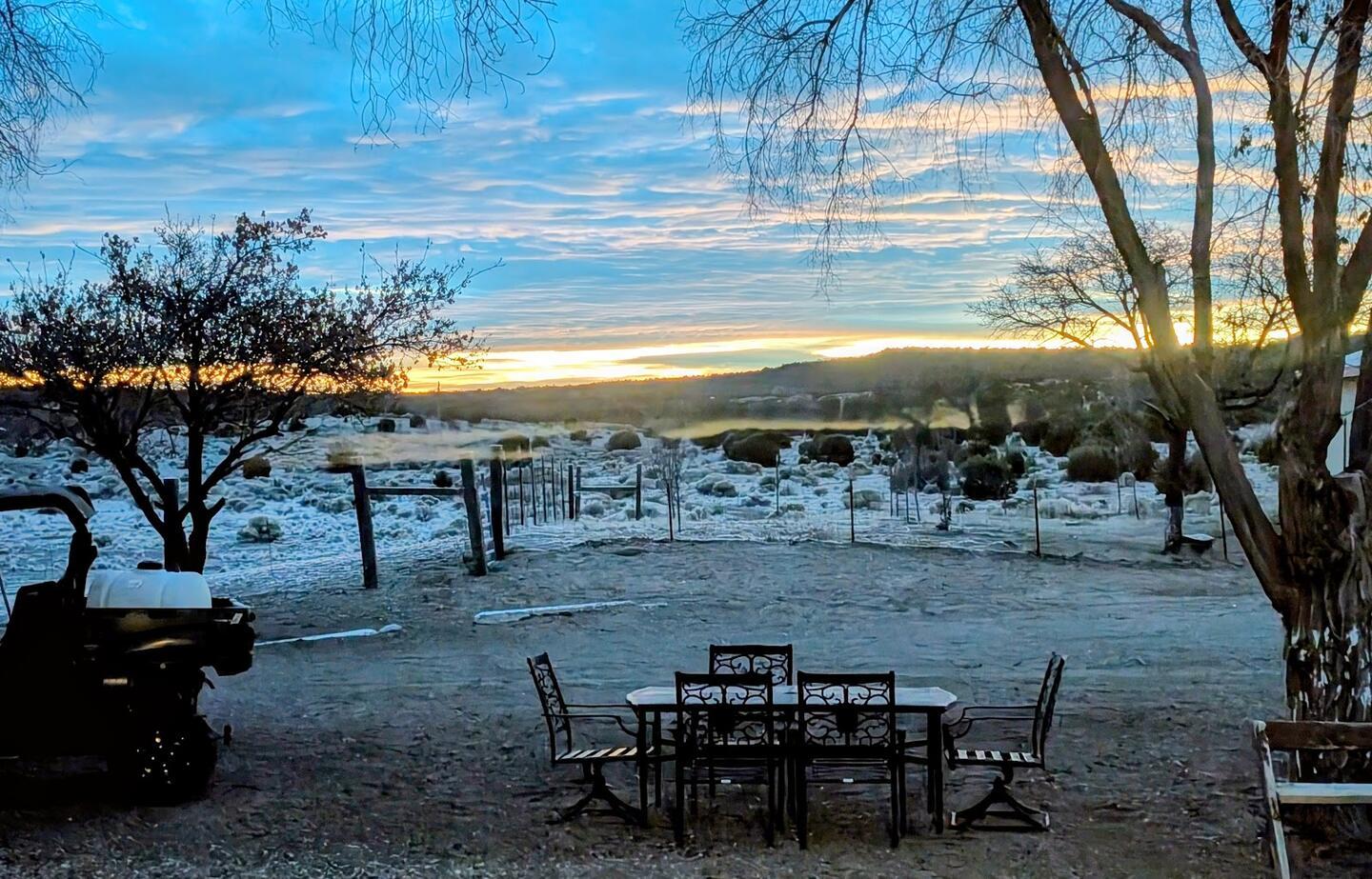 A wrought-iron patio table and chairs on a frost-white yard at sunrise, bare trees framing the pasture and a mobility scooter parked at left