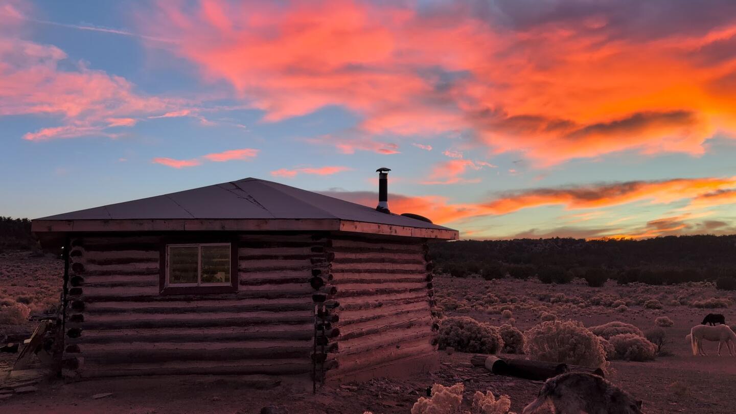 A log cabin with a stovepipe chimney under streaks of orange and pink cloud