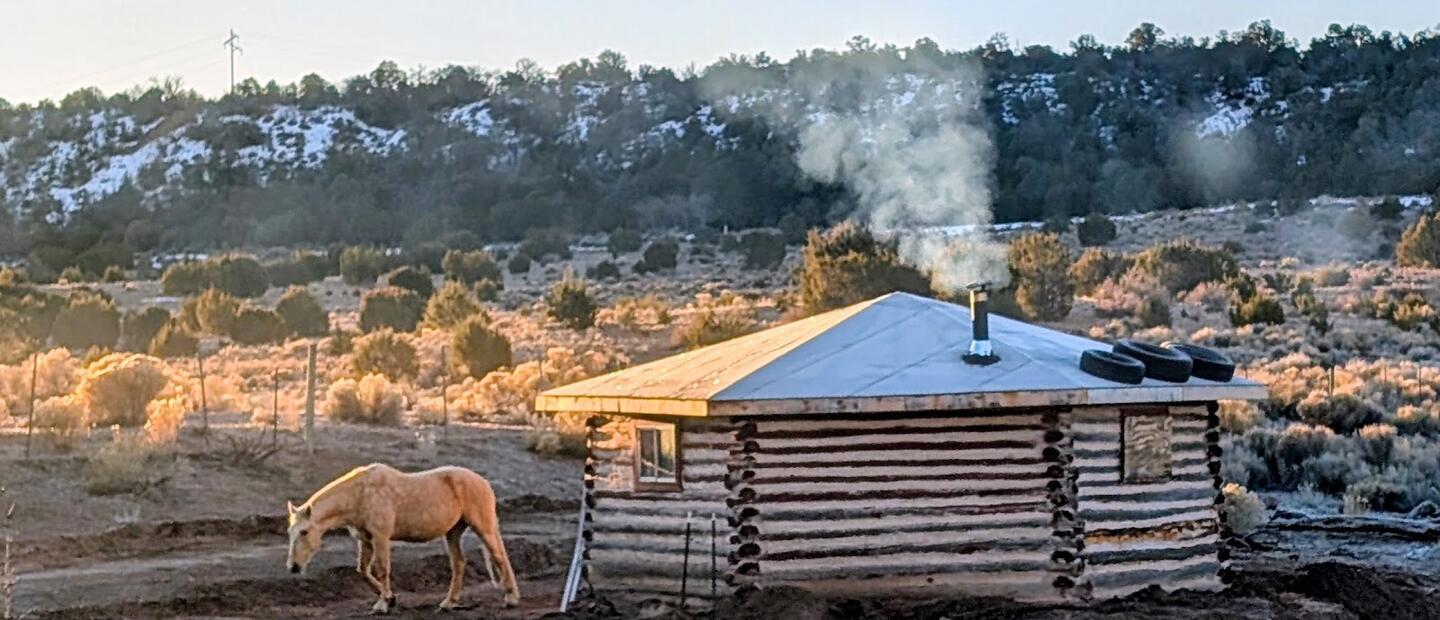 A palomino grazing beside the log cabin as woodsmoke rises from its chimney, snow on the ridge behind