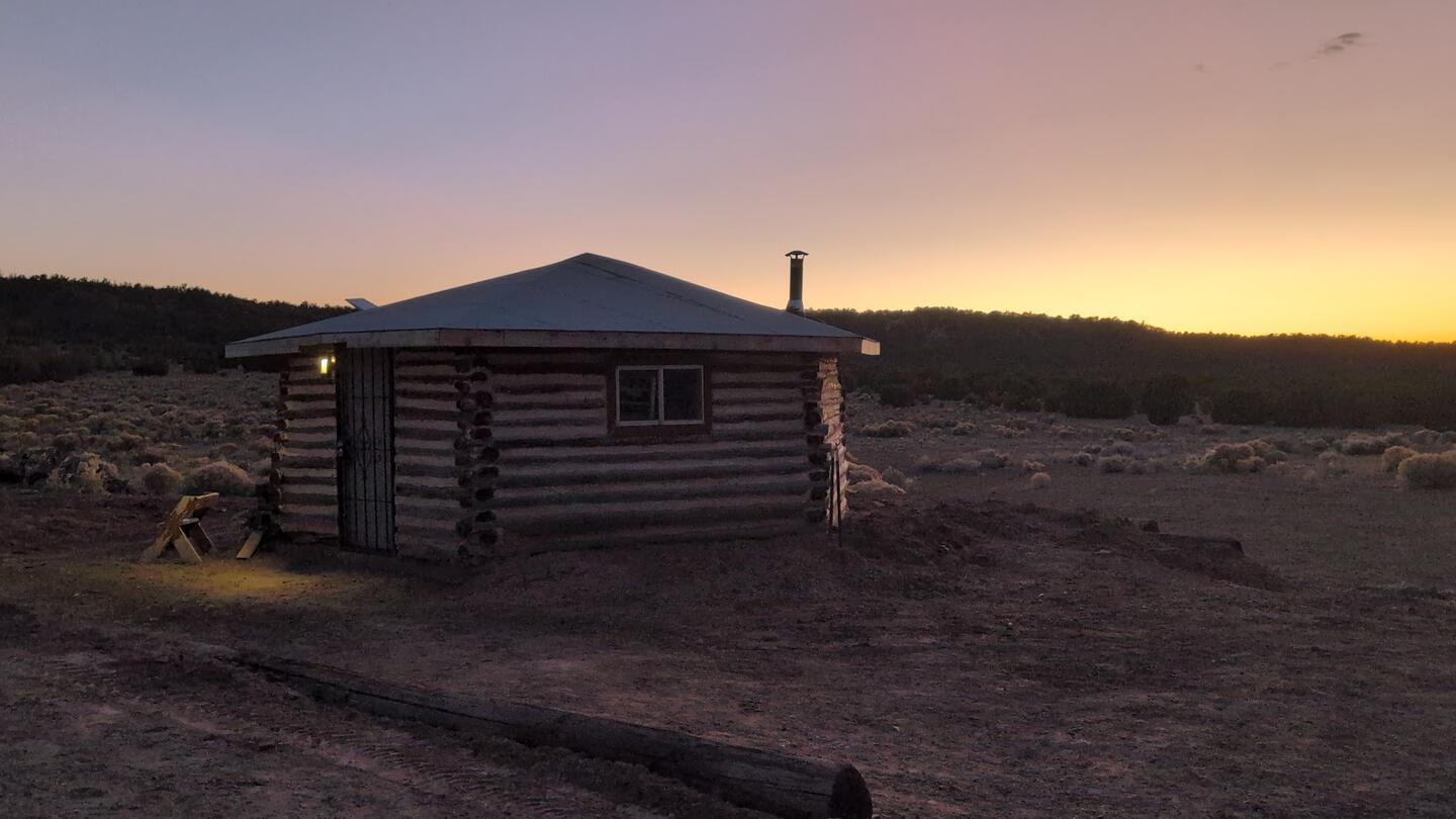 The log cabin at dusk with a lit window, afterglow along the ridge