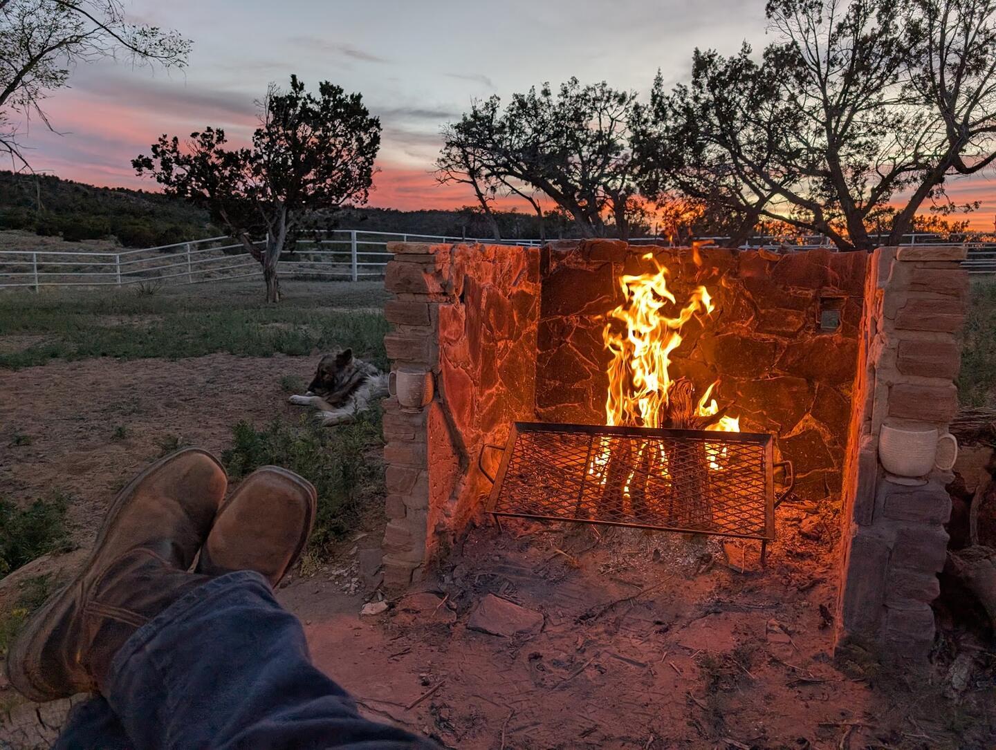 A fire burning in a stone outdoor fireplace at dusk, a pair of boots propped up in front