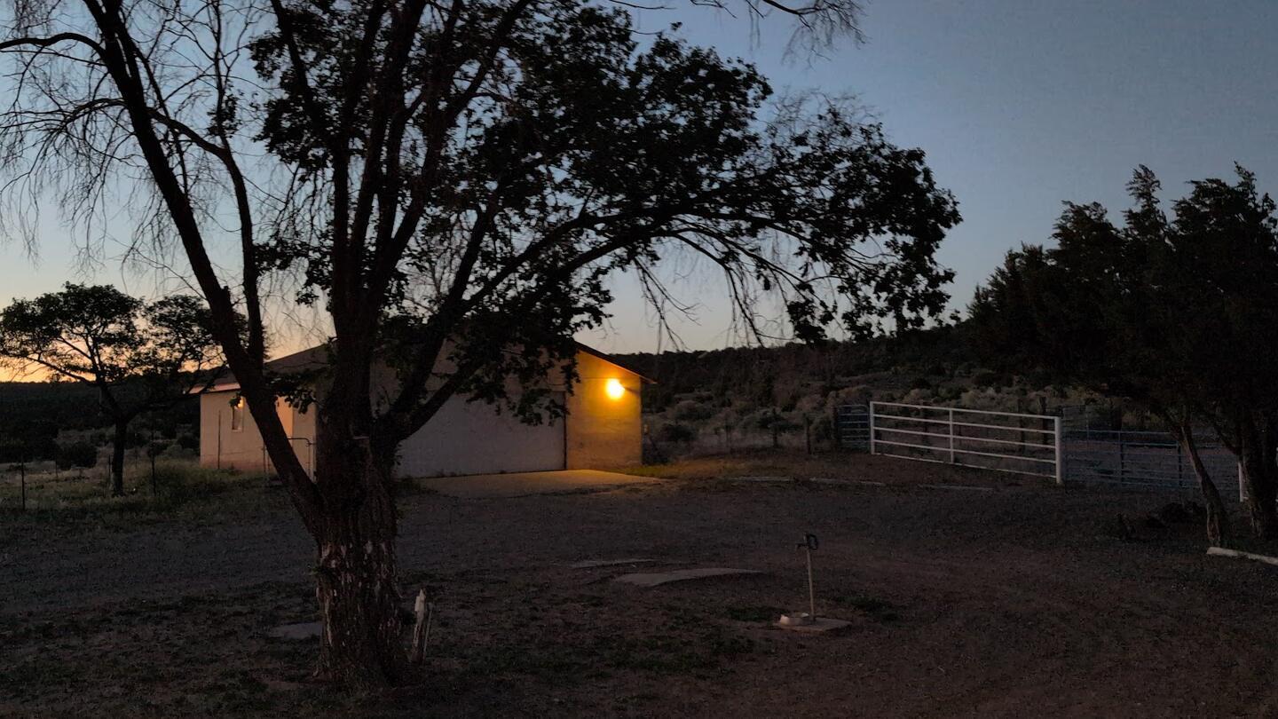 A shed with its porch light on at dusk, juniper and a pipe gate around it
