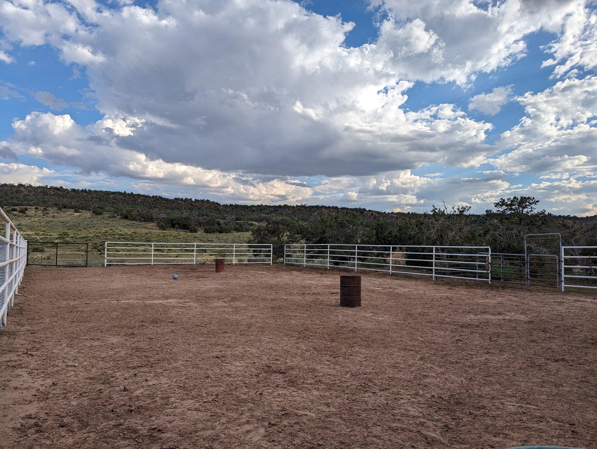 A fenced dirt riding arena with white pipe rails and two barrels, a juniper ridge and tall summer cumulus behind it