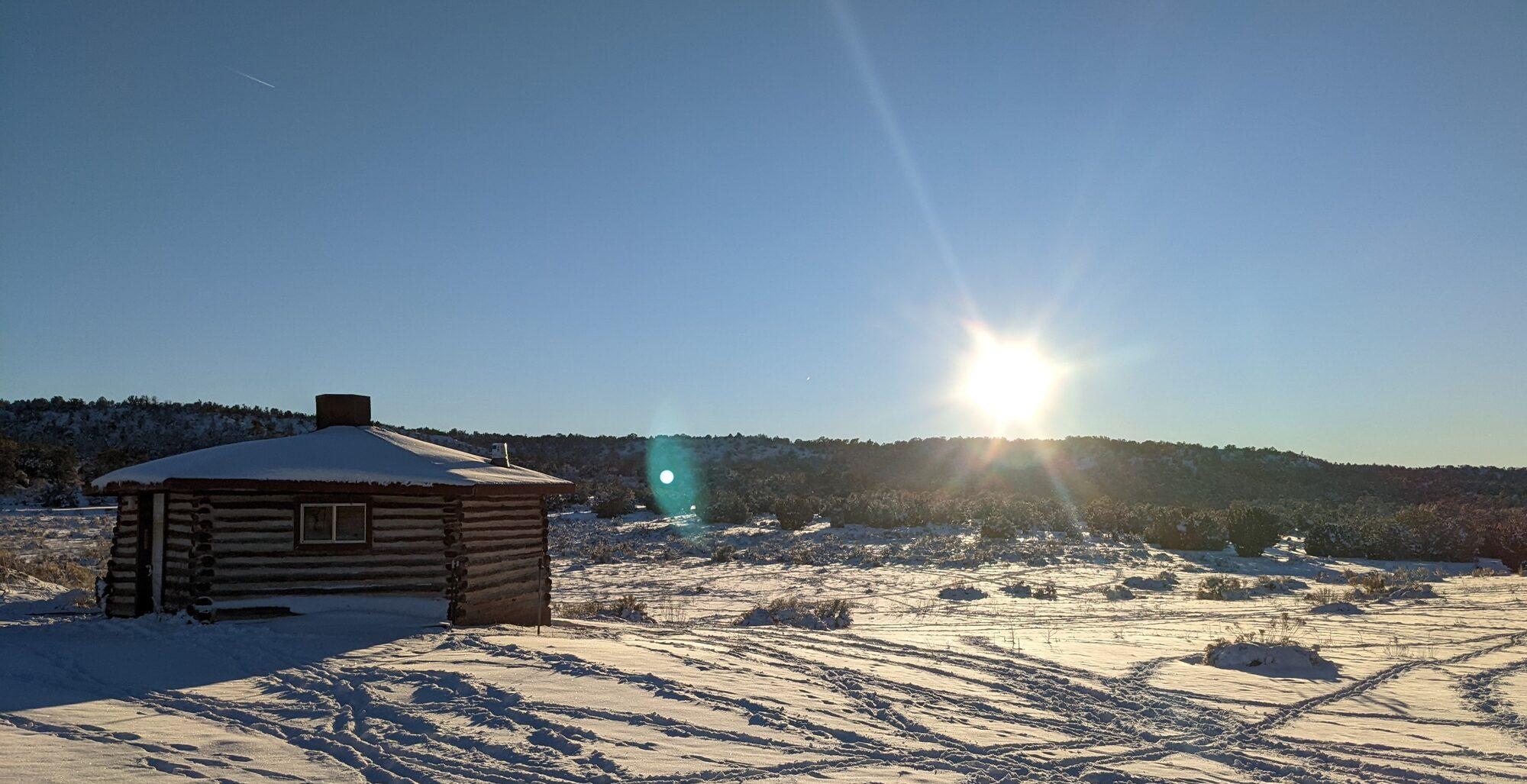 A small log cabin with a stone chimney in fresh snow, low winter sun flaring over the juniper ridge behind it