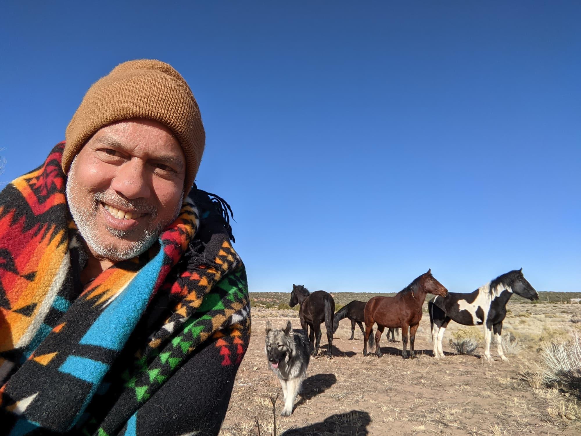 Paul D'Souza wrapped in a brightly patterned wool blanket, with the horse herd and a shepherd dog behind him under a clear blue sky